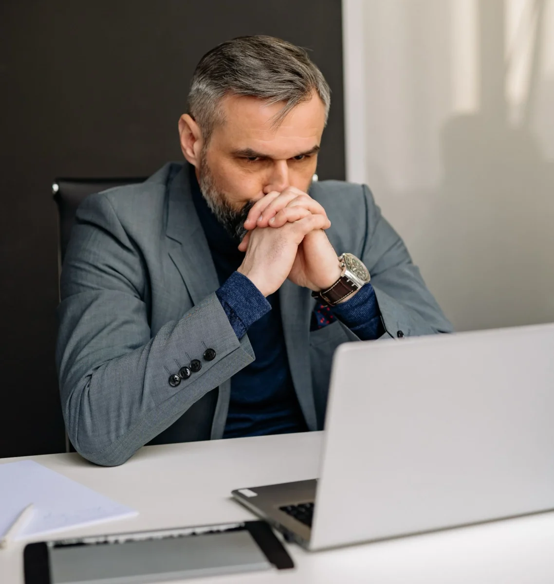 A skeptical middle-aged businessman in a grey suit sitting at an office desk, frowning and staring intently at his laptop screen with his chin resting on his fist.